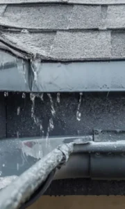 Eau de pluie débordant d'une gouttière abîmée sous un toit en bardeaux bitumés, signe de fuite
